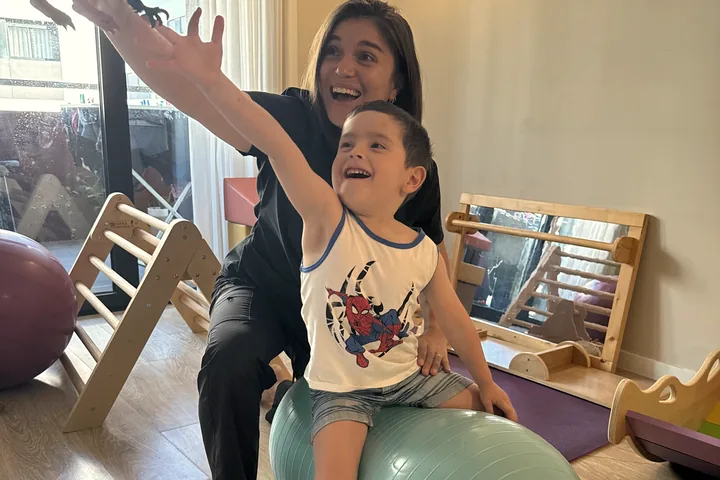 Constanza y un niño sentado en una pelota, los dos riendo mientras alcanzan un juguete.