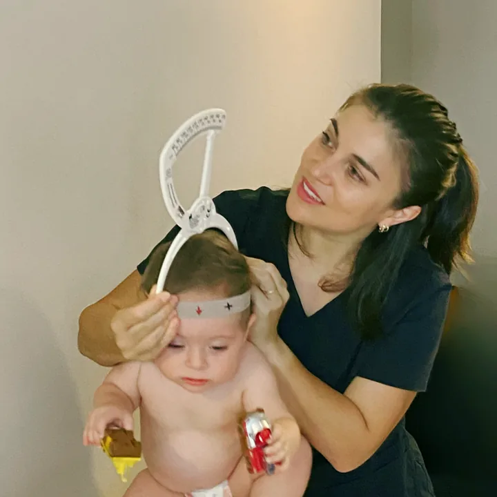 Constanza holds a seated baby and places the craniometer, a white arc with a scale, on the baby's head.